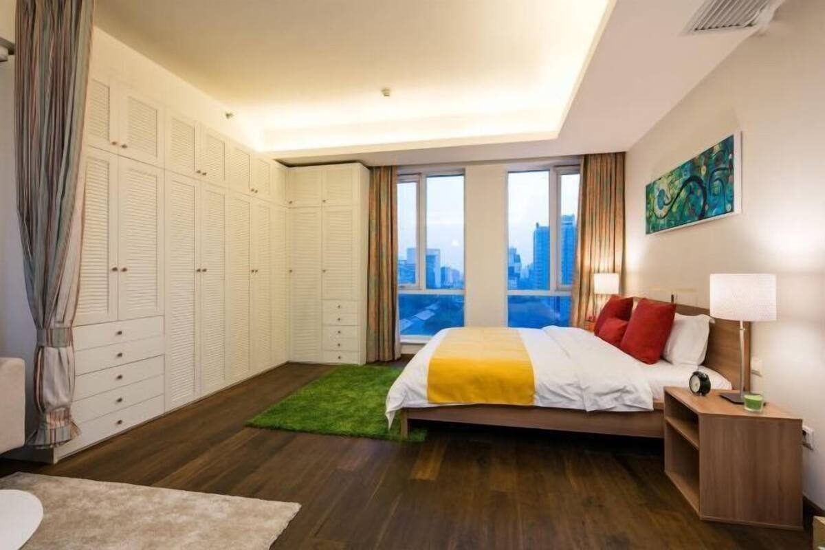 A bedroom with a bed, bedside table, lamp, and a view of the city through large windows.