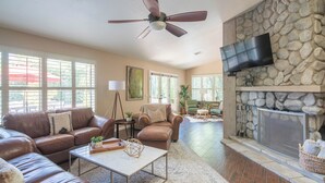A spacious living room with a stone fireplace, leather sofas, a ceiling fan, and large windows. - Scottsdale Treasure: Private Pool and Tranquil Yard Retreat! (Scottsdale)