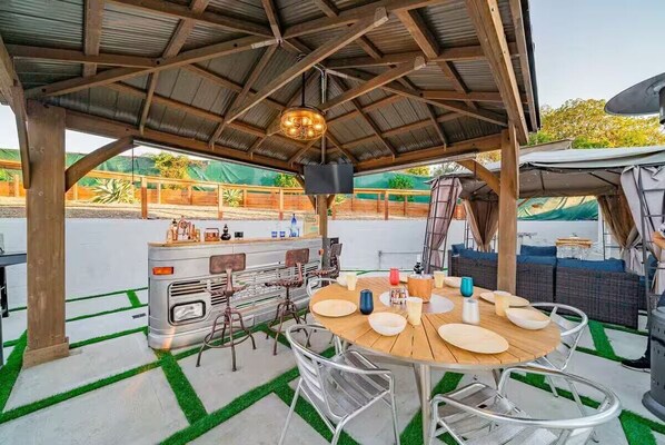Une terrasse ext érieure avec une table ronde en bois et des chaises en métal, un camping-car vintage et un espace abrité avec un toit en métal.