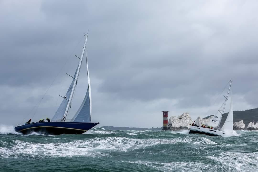 Two sailboats on choppy waters with a rocky coastline and a lighthouse in the background.