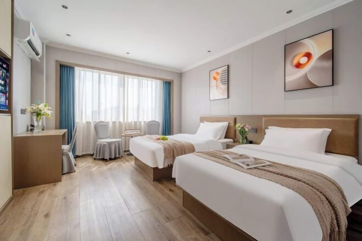 Habitación de hotel con dos camas, un escritorio, una silla, una ventana con cortinas y obras de arte en las paredes.