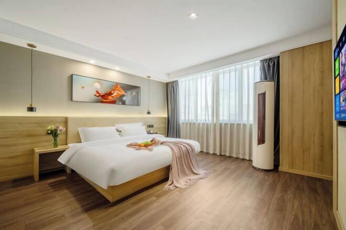 Una habitación de hotel moderna con una cama grande, una mesita de noche con un florero, un cuadro en la pared y un ventanal con cortinas.