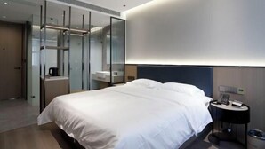 A modern hotel room with a large bed, a bedside table with a phone and a lamp, a glass-enclosed walk-in closet, and a wall-mounted light fixture.