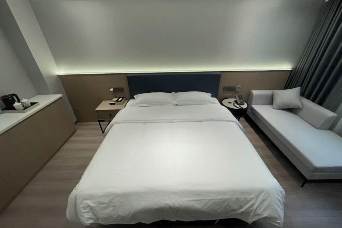 A modern hotel room with a bed, a bedside table, a lamp, a sofa, and a curtain.