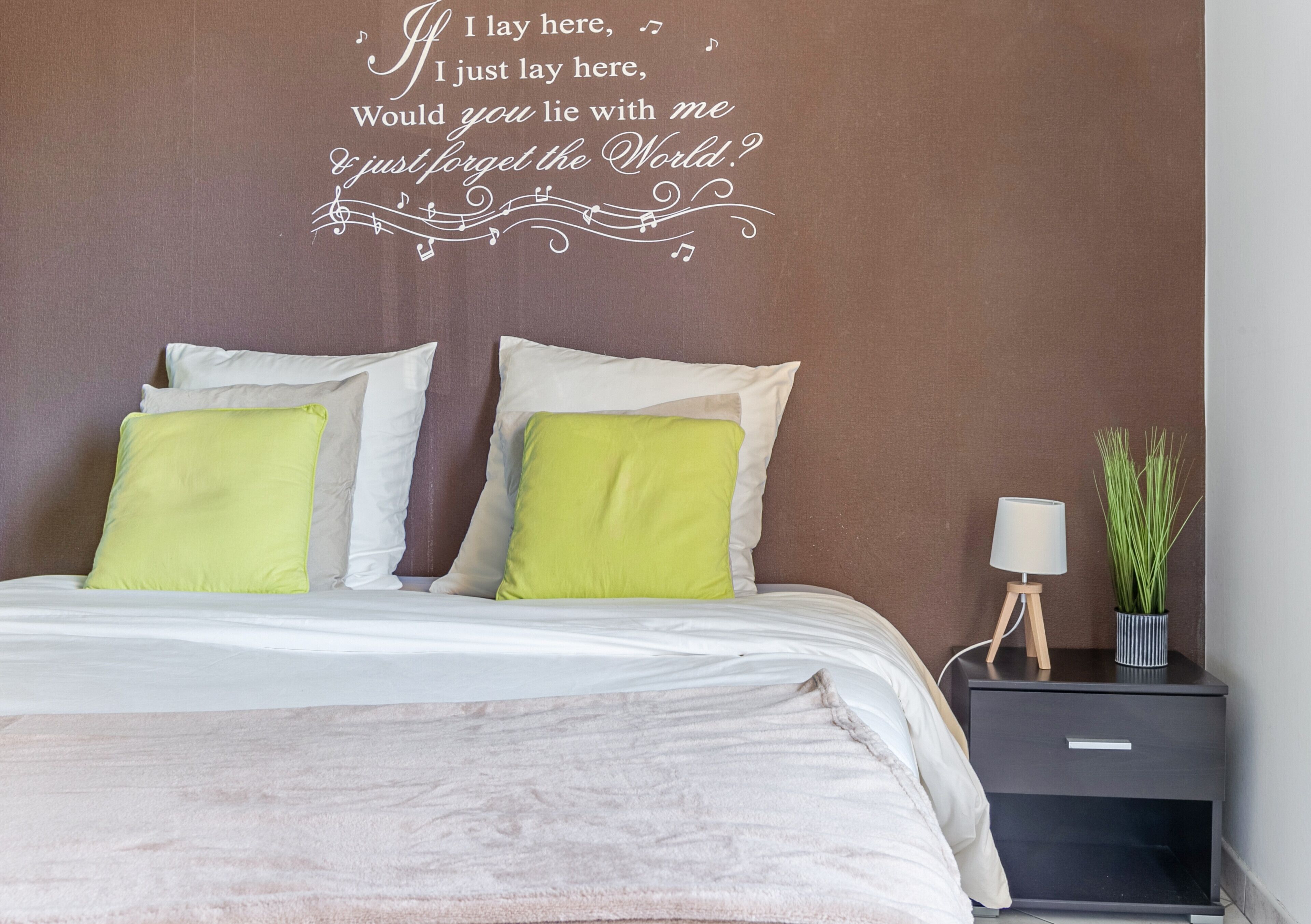 Une chambre à coucher avec un lit, des oreillers, une table de chevet, une lampe et un mur orné d’une citation décorative.