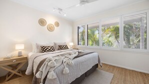Une chambre à coucher comprenant un lit, des tables de chevet, une fenêtre avec des stores et un ventilateur de plafond.