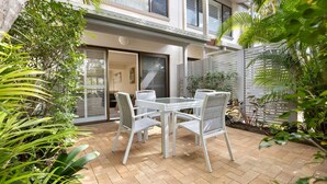Une terrasse avec une table et des chaises, entourée de verdure et d’une clôture blanche.