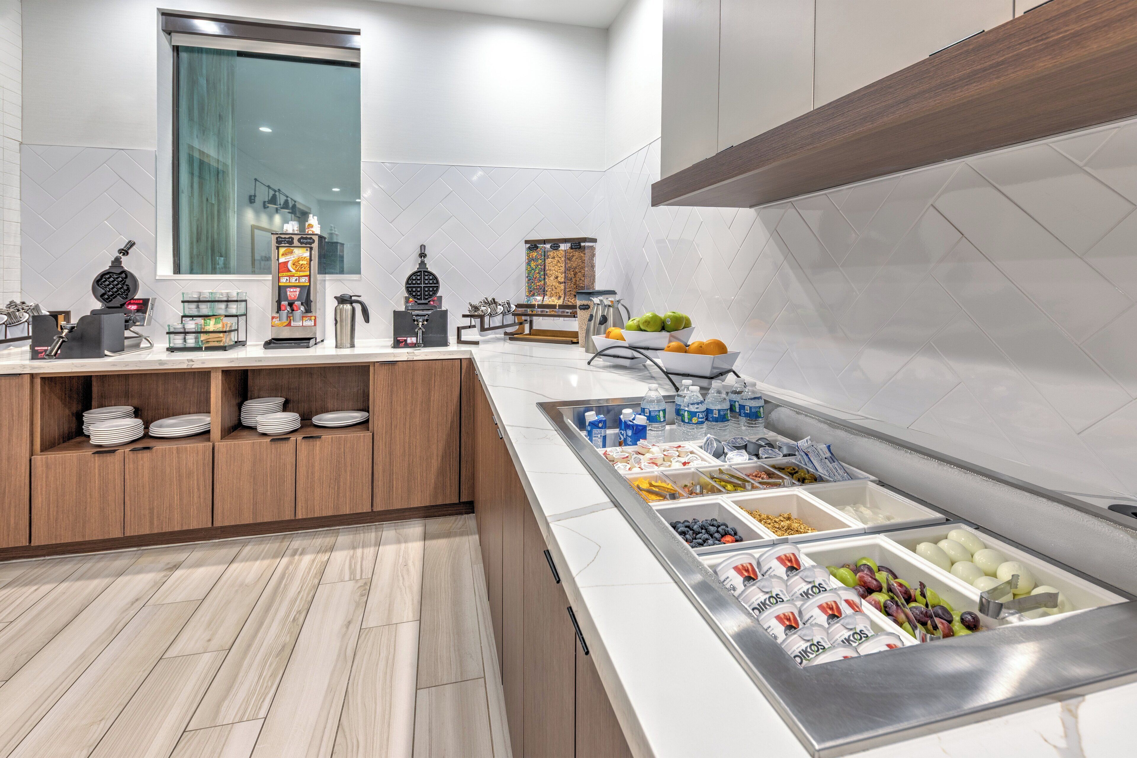Cocina moderna con barra desayunadora, paredes de azulejos blancos, armarios de madera y estación para desayunar de acero inoxidable con diferentes alimentos.