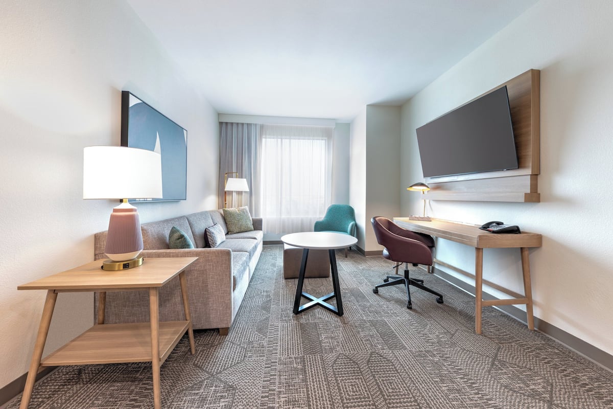 A hotel room with a sofa, a round table, a desk, a chair, a TV mounted on the wall, and a lamp on the side table.