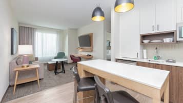 A modern hotel room with a dining area, kitchenette, and a seating area with a sofa and chairs.