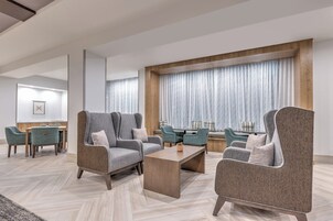 Un hall de hotel moderno con sillones tapizados grises, una mesa de centro de madera y un amplio ventanal con cortinas de gasa.