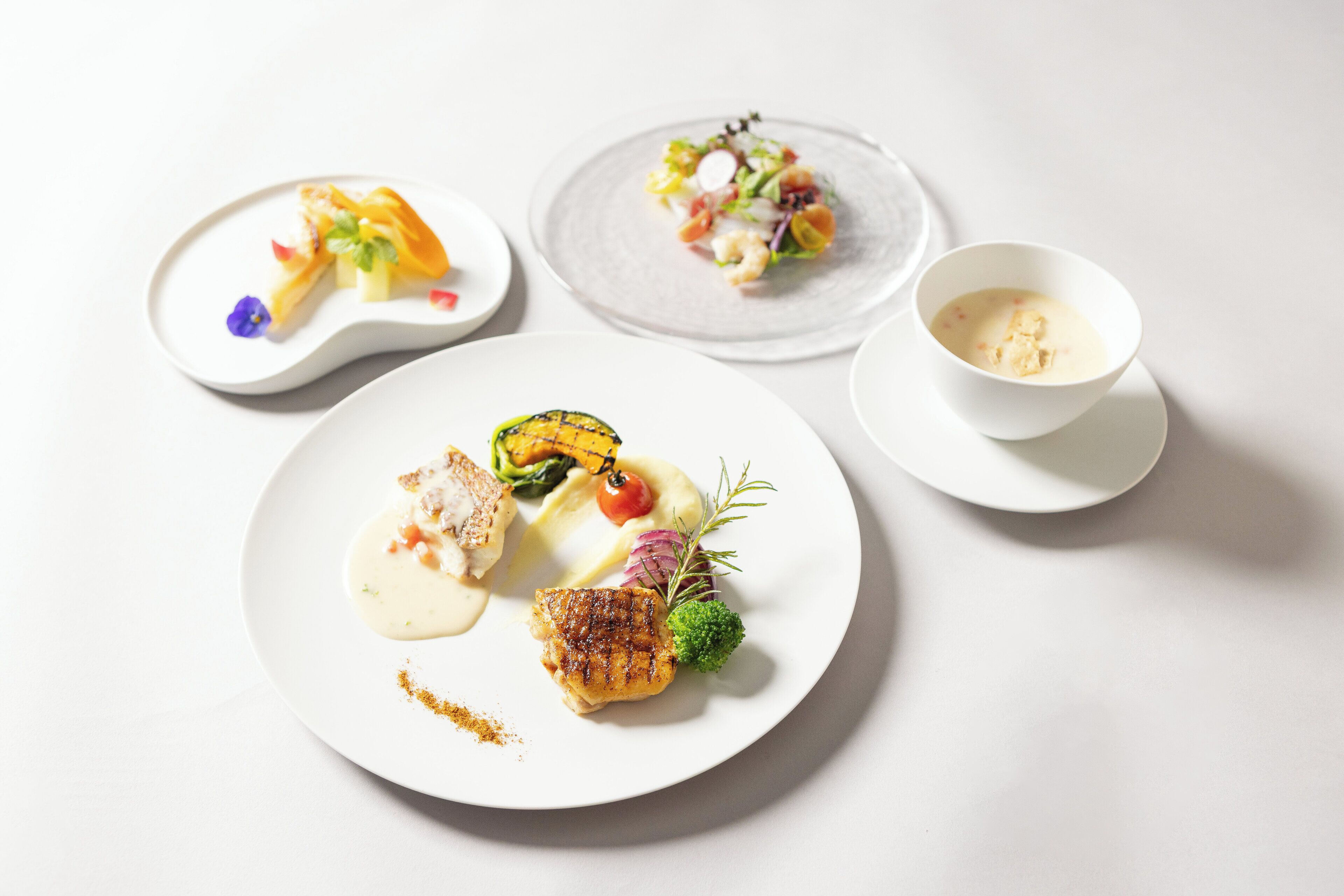 Three plates of food with a bowl of soup on a white table.