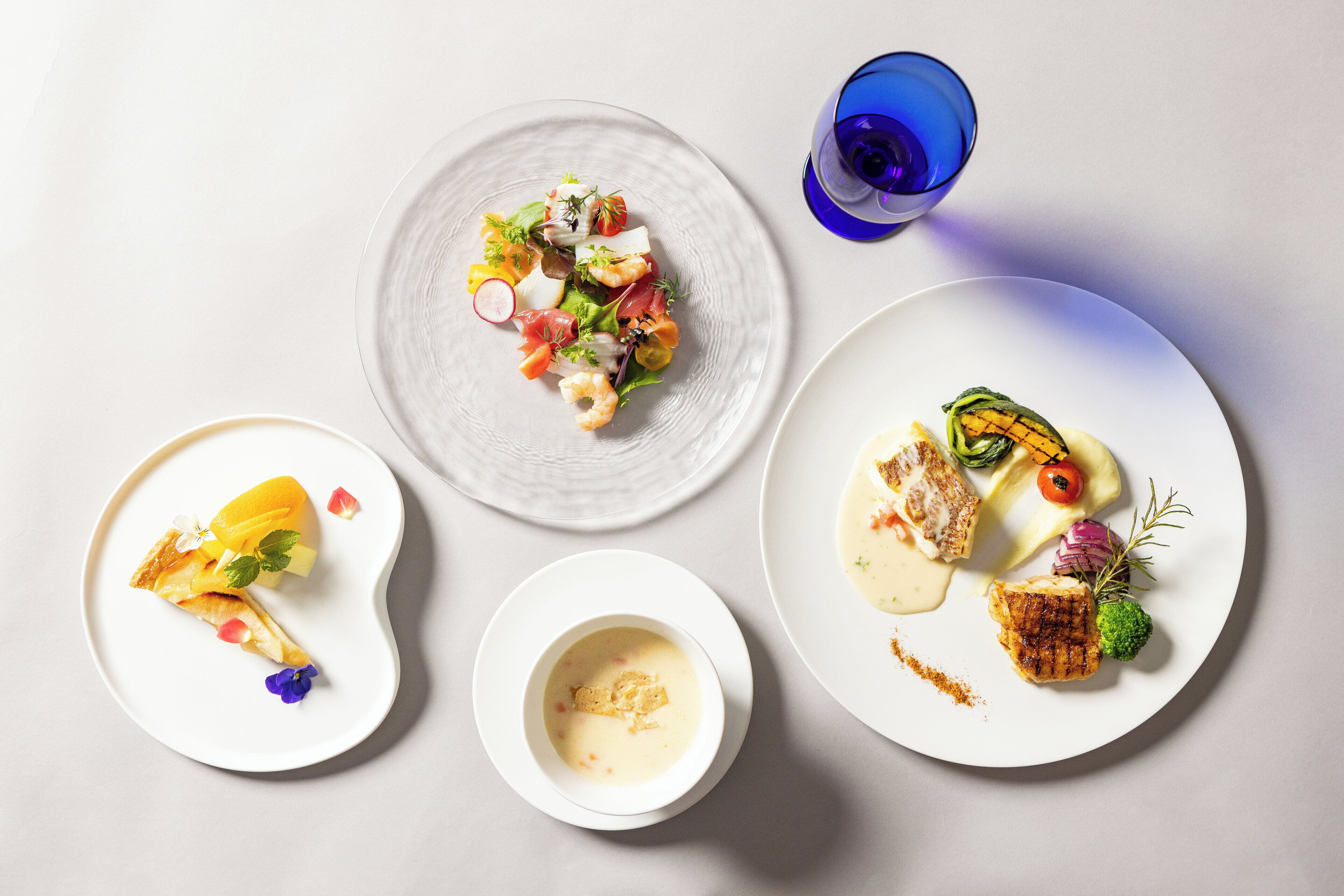 A table with three plates of food, a glass of blue liquid, and a small bowl of soup.