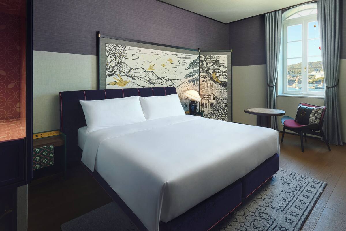 A neatly made bed in a hotel room with a large window, a small table, and a chair.