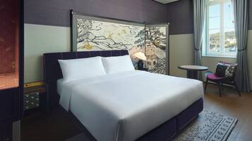 A neatly made bed in a hotel room with a large window, a small table, and a chair.