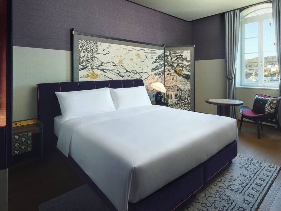 A neatly made bed in a hotel room with a large window, a small table, and a chair.