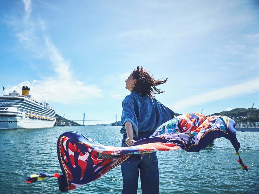 A person holding a colorful kite by the water with a cruise ship in the background.