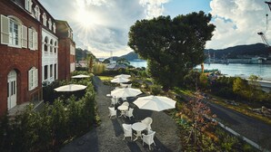 A paved outdoor area with white tables and umbrellas, a brick building with arched windows, and a river with a bridge in the background. - Hotel Indigo Nagasaki Glover Street by IHG (Nagasaki)