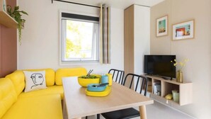 A modern living space with a yellow sofa, a dining table, a TV, and a window with curtains. - Camping 3 étoiles - Piscine  - eehdba (Portel-des-Corbières)