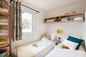Une chambre avec deux lits, une fenêtre avec des rideaux, une étagère remplie de livres et un petit miroir rond.