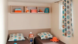 A room with two single beds, a shelf with storage boxes, a clock, and a window with a floral curtain. - ZEcamping 3 étoiles piscine chauffée mobil-home 25m2 4 pers (Portel-des-Corbières)