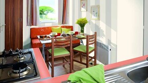 A compact kitchen with a gas stove, wooden chairs, a dining table, and a window with curtains. - ZEcamping 3 étoiles piscine chauffée mobil-home 25m2 4 pers (Portel-des-Corbières)