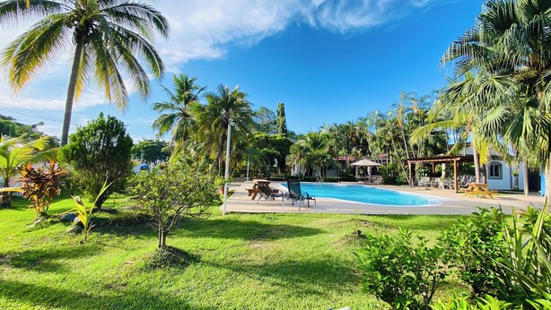 Un área de piscina con sillones reclinables y un pabellón, rodeada de palmeras y follaje.