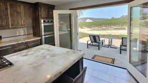 A kitchen with a marble countertop, double ovens, and wooden cabinets. There is a sliding glass door leading to an outdoor patio with a table and chairs.