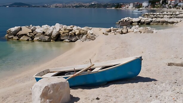 A beach with a small boat, rocky shore, and buildings in the background. - Adriatic Split beach apartment with garden (Podstrana)