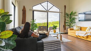 Una sala de estar moderna con un amplio ventanal corredera que da a un balcón, un sofá, una mesa de centro y un televisor montado en la pared.