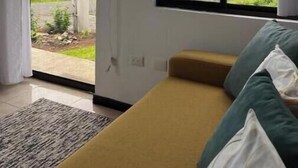Habitación con un ventanal grande que ofrece vistas a un paisaje verde y un edificio lejano. Hay una cama con almohada verde y blanca, un ventilador blanco en la pared y una pequeña alfombra en el suelo.
