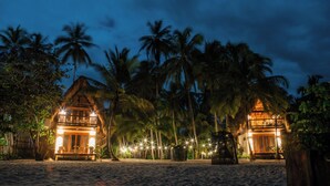 Two thatched-roof buildings with illuminated interiors, surrounded by palm trees on a sandy beach at night. - GAELIA BEACH (Santa Marta)