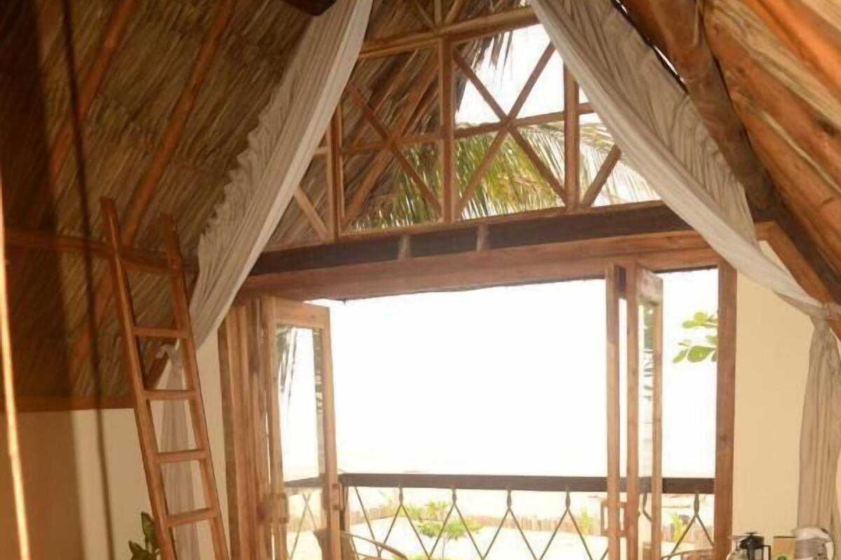 A wooden room with a bed, a ladder, a small table, and a view of the beach.
