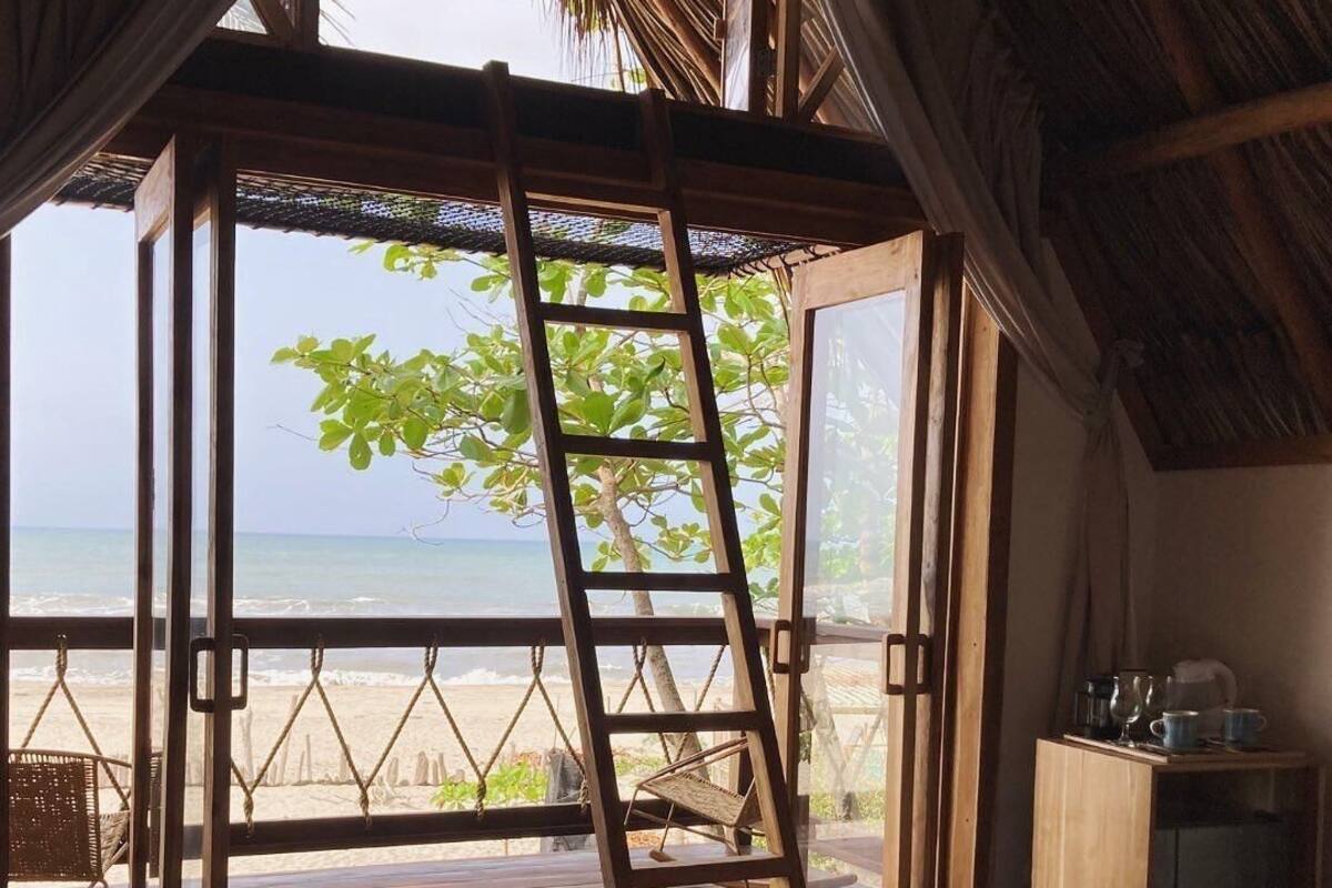 A wooden ladder in a room with a view of the beach, a small table with cups, and a wooden cabinet.