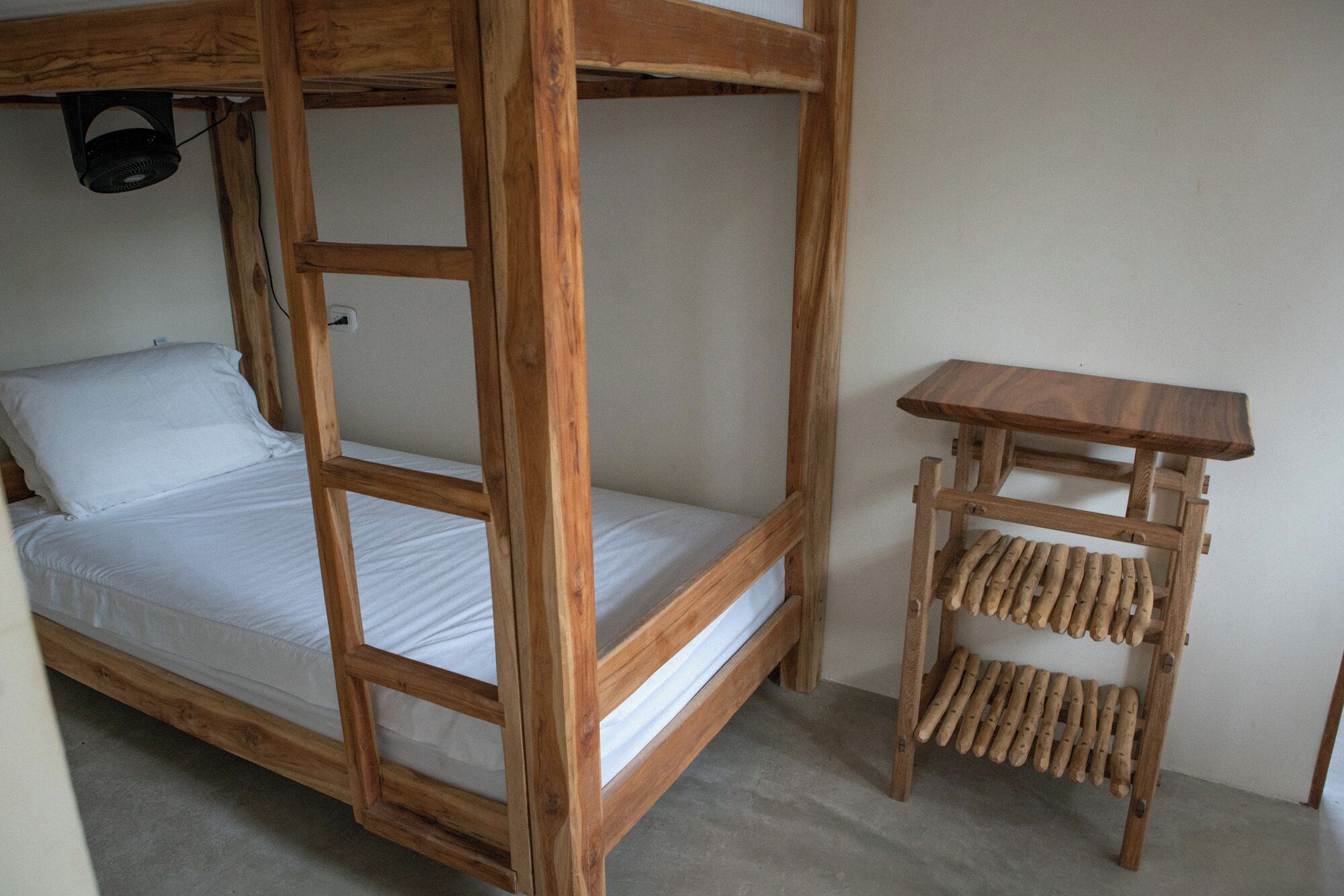 Een stapelbed met houten frame en witte beddengoed, een klein houten tafeltje met planken, en een plafondventilator.