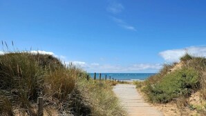 A sandy pathway leading to a beach with dunes and a clear blue sky.
