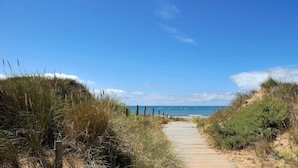 A sandy pathway leading to a beach with dunes and a clear blue sky.