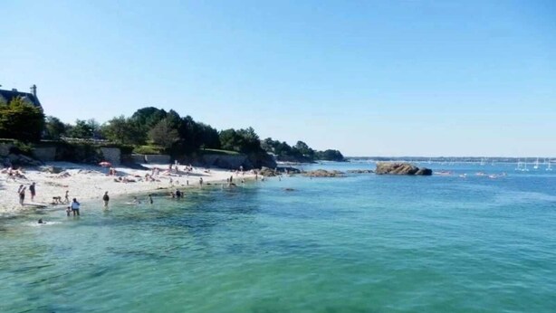 Una playa con agua cristalina, gente nadando y un promontorio rocoso en la distancia.