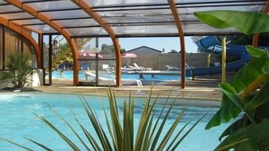 A covered swimming pool with a transparent roof, surrounded by lounge chairs and a water slide.