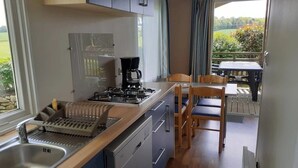 A compact kitchen with a sink, stove, and coffee maker. There is a dining area with a table and chairs. - ZEcamping 3 étoiles toboggans mobil-home 37m2 4 pers (Inguiniel)