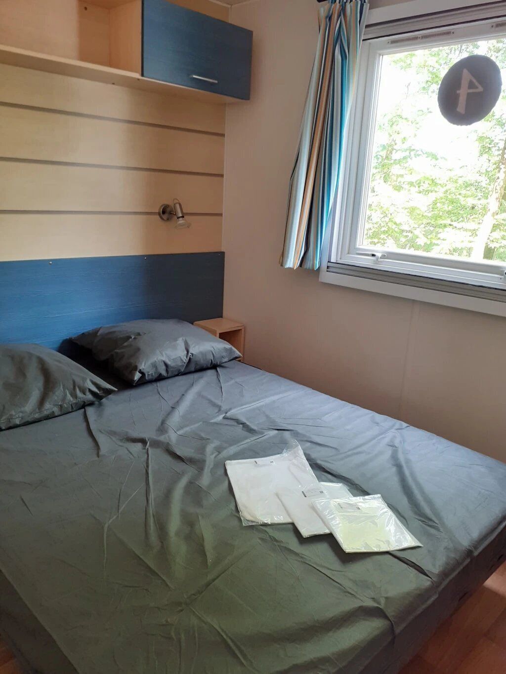 A single bed with a grey mattress and pillows, a wooden shelf above, a small bedside table, and a window with curtains.