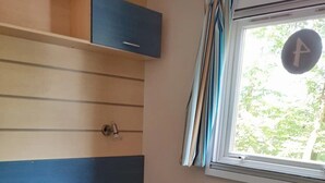 A single bed with a grey mattress and pillows, a wooden shelf above, a small bedside table, and a window with curtains. - ZEcamping 3 étoiles toboggans mobil-home 37m2 4 pers (Inguiniel)