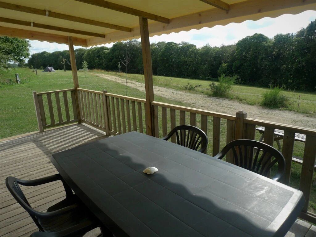 Une terrasse extérieure en bois, dotée d’une table et de chaises, entourée d’une balustrade en bois, et offrant une vue sur une zone herbeuse avec des arbres et une tente visible au loin.