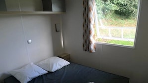 Een enkel bed met een donkere matras en witte kussens, een klein nachtkastje, een raam met gordijnen en uitzicht op groen buiten.