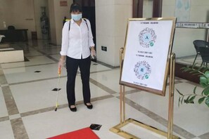 Une personne se tient à côté d’un tableau blanc sur lequel sont écrits du texte et des symboles, dans un hall d’entrée doté d’un tapis rouge et d’un sol à carreaux.