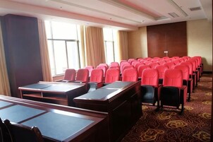 Un salón de conferencias con filas de sillas rojas y mesas, ventanas grandes con cortinas y una alfombra estampada.