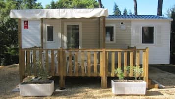 Un piccolo camper a un solo piano con una veranda in legno e due vasi con piante.