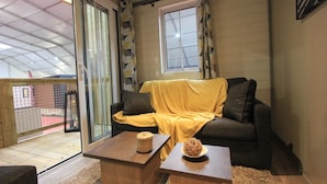 A cozy living room with a sofa covered in a yellow blanket, two wooden side tables with decorative items, and a view of a balcony through a glass door.