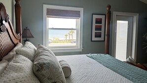 Un dormitorio con un ventanal grande, una cama con muchas almohadas, una mesita de noche con una lámpara y un cuadro enmarcado en la pared.