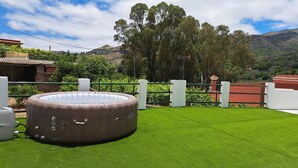 A circular outdoor hot tub on artificial grass, surrounded by a white fence, with a mountainous landscape in the background.